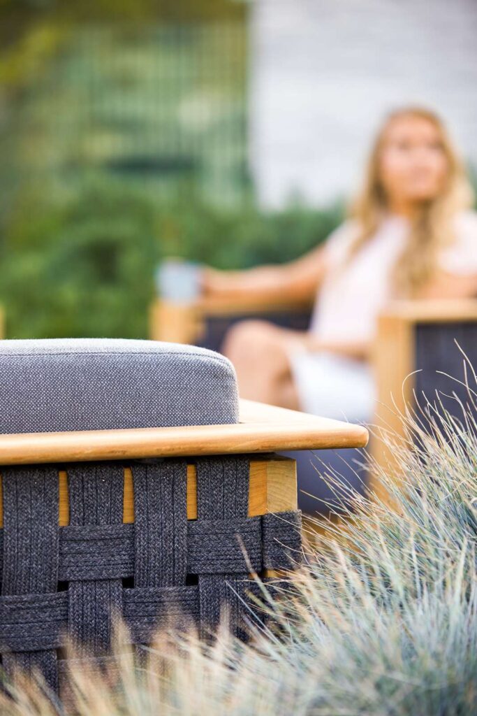 zbliżenie na fragment fotela z widoczną plecionką i ramą z drewna teak, w tle rozmyta postać kobiety