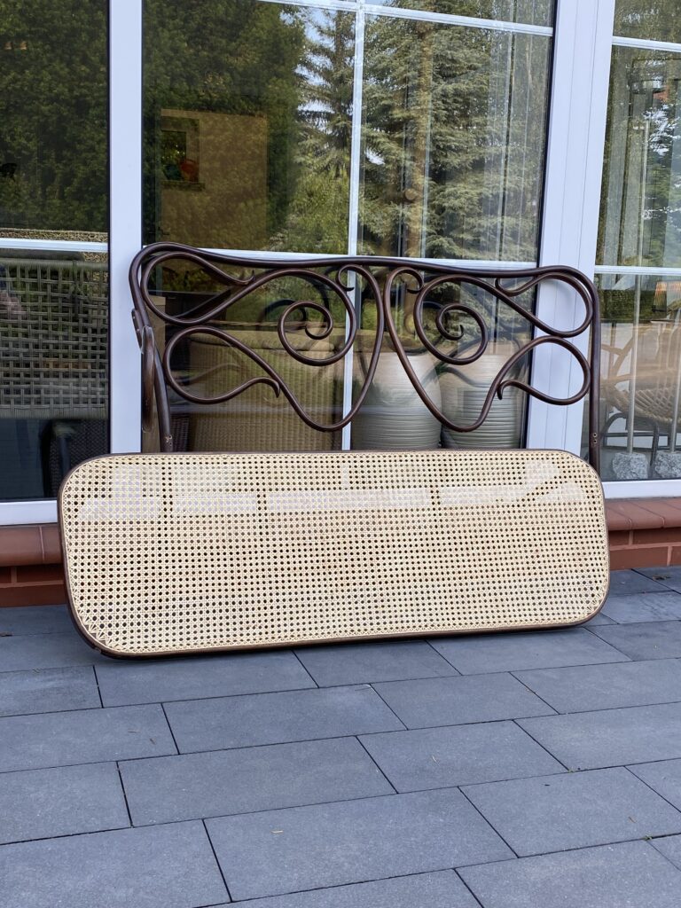 Widok na antyczną sofę z siedziskiem ręcznie wyplatanym rattanem. Zdjęcie zrobione na tle szyby z odbijającymi się drzewami. Siedzisko sofy jest postawione przy ramie i oparciu mebla.