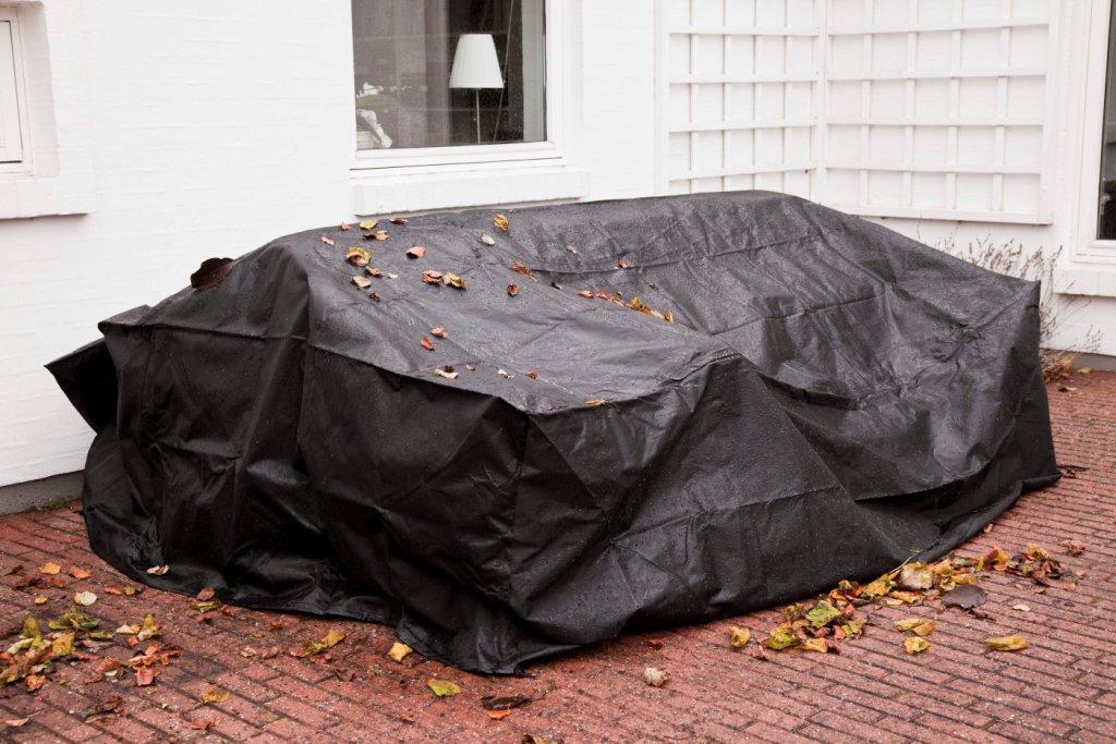 Grupa mebli ogrodowych pod pokrowcem. Kolor ciemno szary. Na zdjęciu widoczne spadające liście drzew