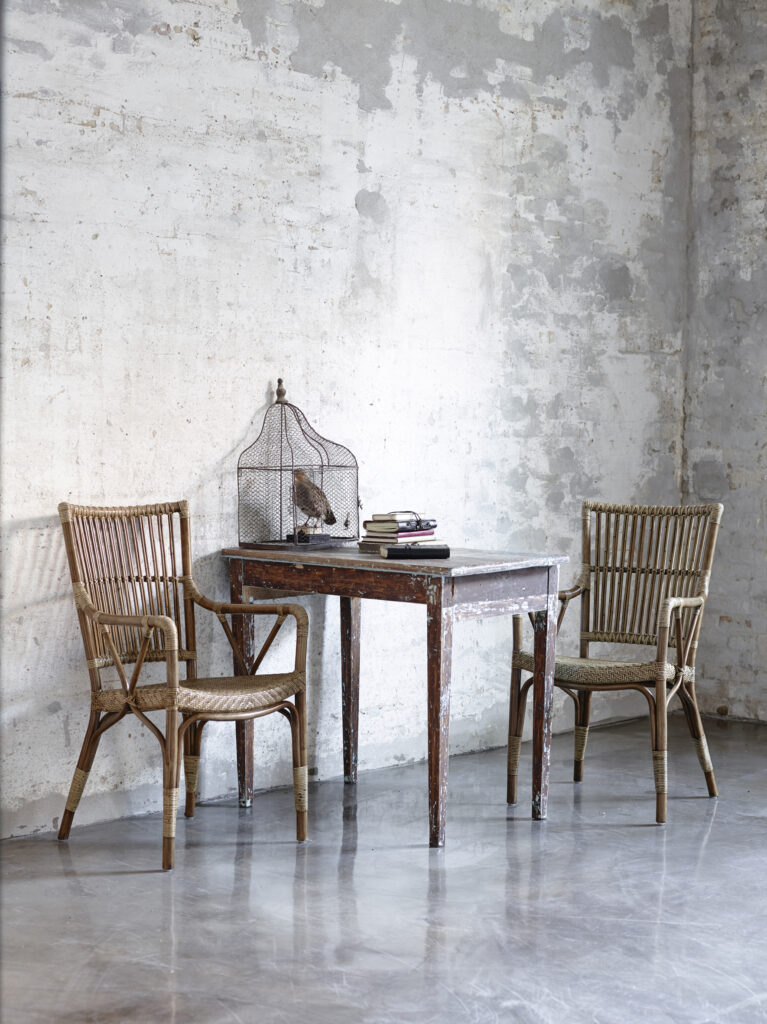 widok na rustykalne pomieszczeni, przy starym, kwadratowym stoliku stoją dwa rattanowe krzesła z podłokietnikami w kolorze beżowym