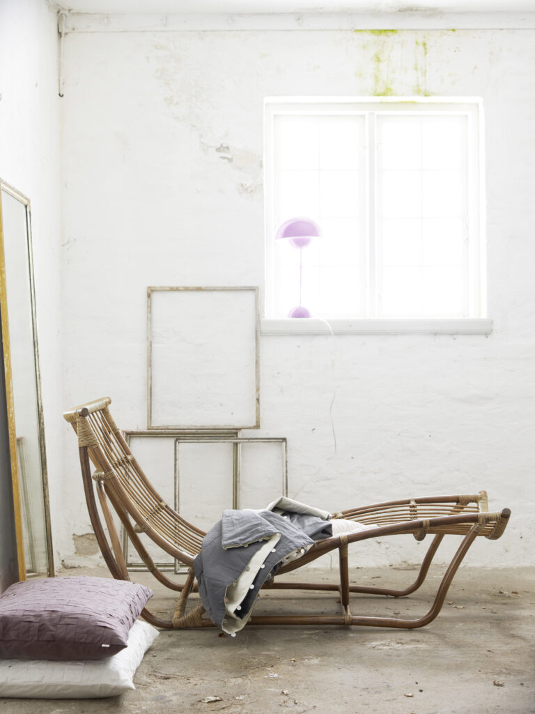 w rustykalnym pomieszczeniu stoi leżanka z rattanu w formie ażurowego leżaka, na leżance leży narzuta do przykrycia, leżanka w kolorze miodowym
