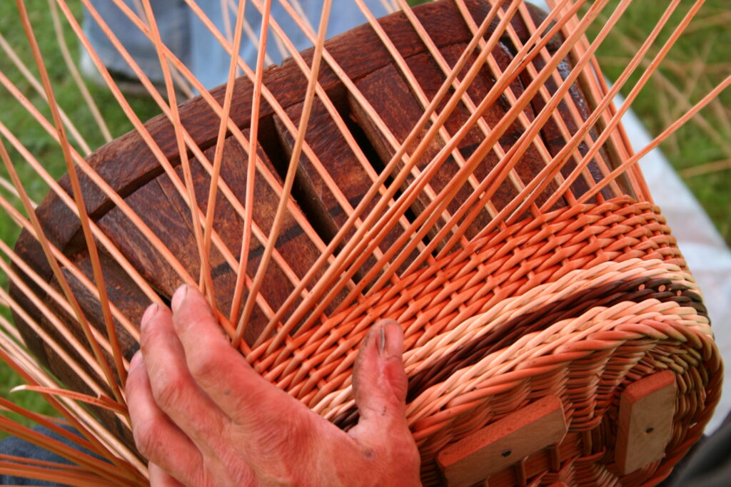 Zbliżenie na wyplatany koszyk z wikliny, wypleciony fragment kosza z kolorową wstawką (kolor biały i ciemno brązowy), widać rękę plecionkarza