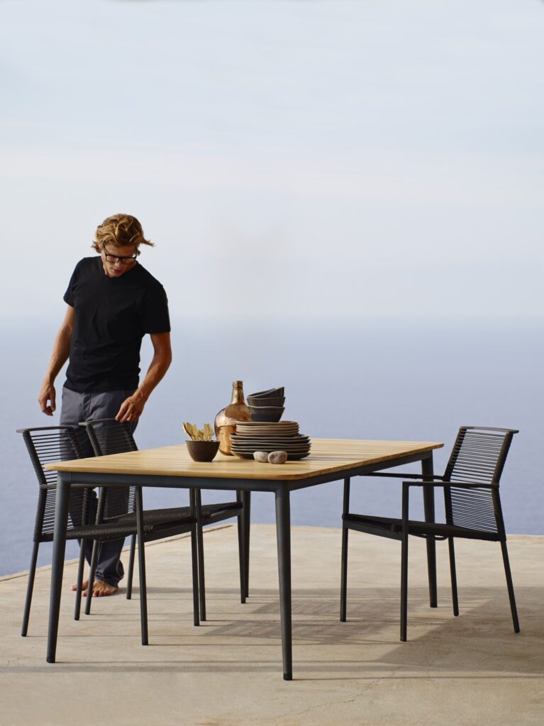 dwa krzesła strunowe stoją przy stole z blatem teakowym, wszystko wygląda bardzo subtelnie, przy stole młody mężczyzna