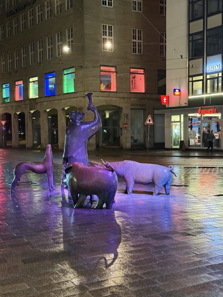 Rzeźba świniopasa i stada świń na ulicy Sögestraße w Bremie po deszczu, na tle kolorowo oświetlonych budynków