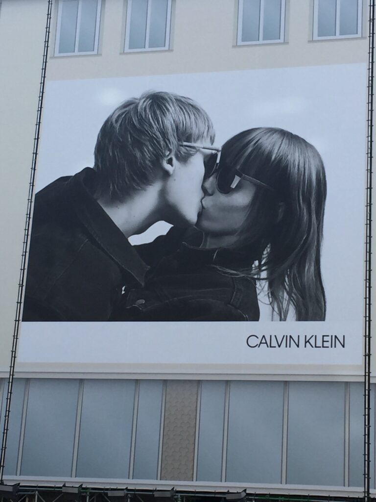 Czarno-biała wielkoformatowa reklama Calvin Klein przedstawiająca całującą się parę na elewacji budynku w przestrzeni miejskiej.