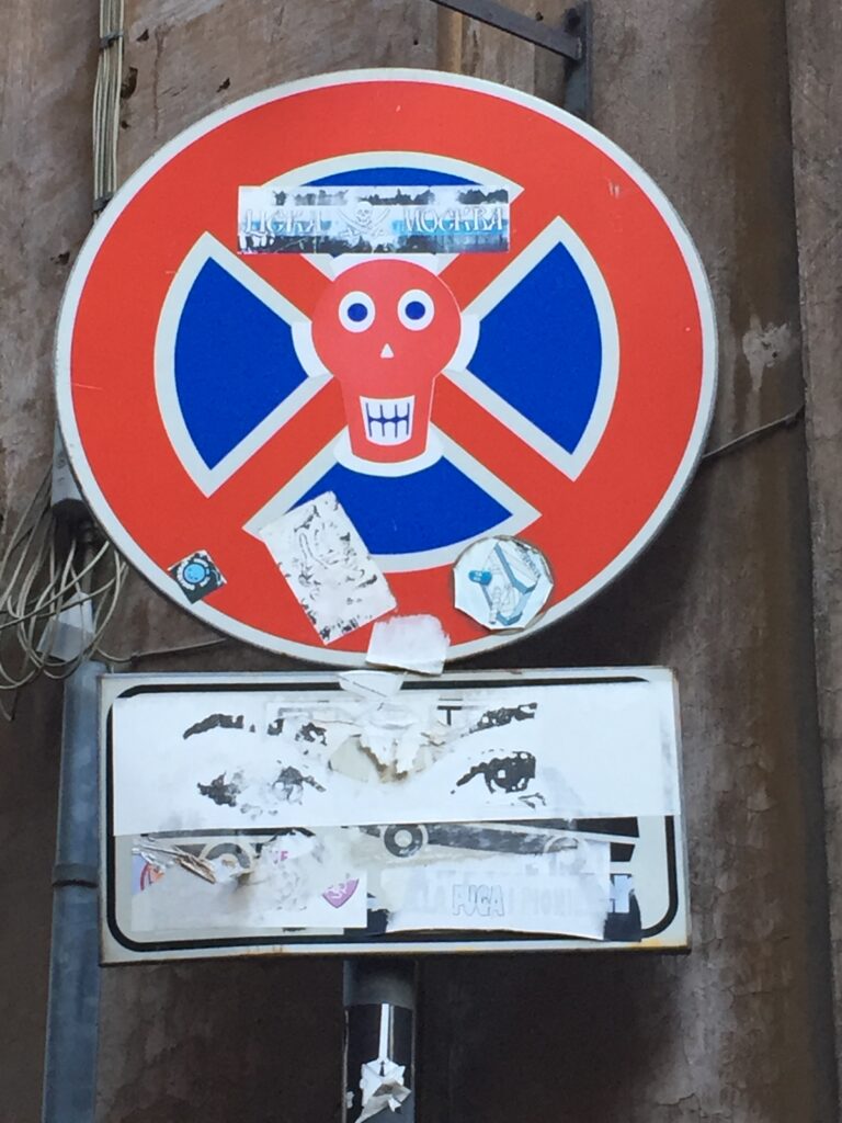 Znak drogowy w Rzymie pokryty naklejkami i śladami plakatów, ukazujący warstwowość miejskiej przestrzeni i kulturę uliczną.